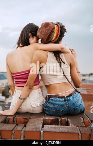 Vista posteriore di una giovane donna che abbraccia un'amica che indossa un cappello a maglia mentre si siede sul tetto Foto Stock