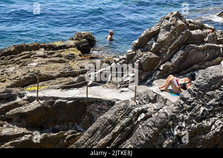 Una donna che legge sulla scogliera e un uomo che nuota sulla riva del mare della passeggiata Anita Garibaldi, meta turistica popolare a Genova, Nervi, Liguria Foto Stock