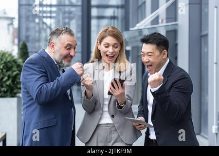 un gruppo di colleghi, i dipendenti, leggono buone notizie guardando lo smartphone. Il team felice si rallegra dell'investimento di successo durante l'utilizzo e la navigazione sul telefono cellulare. In piedi all'esterno vicino all'edificio degli uffici. Foto Stock