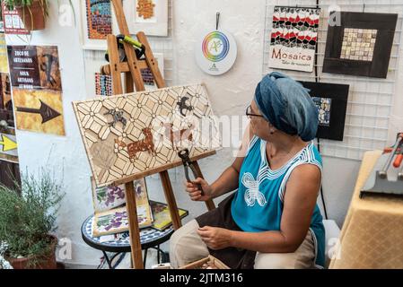 Frigiliana, Malaga, 27 agosto 2022: Artigiano che lavora su una piastrella a mosaico con rondini e cani che lavorano su una strada a Frigiliana in occasione di Foto Stock