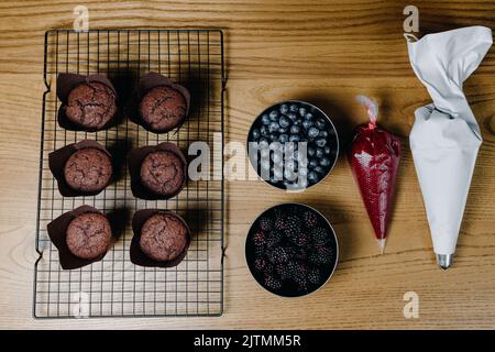 Cupcake al cioccolato appena sfornati con ingredienti nelle vicinanze. Decorazione con crema e frutti di bosco freschi Foto Stock