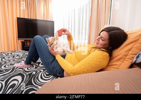Bella giovane donna ispanica sorridente vestita di giacca gialla, sdraiata sul suo letto giocando con il suo gattino con una palla rossa di filato Foto Stock