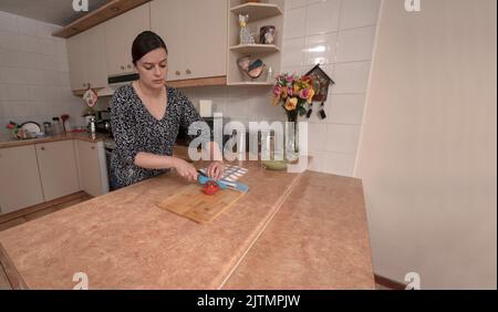 Bella giovane donna ispanica affettare un pomodoro con un coltello blu su un asse di legno al suo bancone da cucina Foto Stock