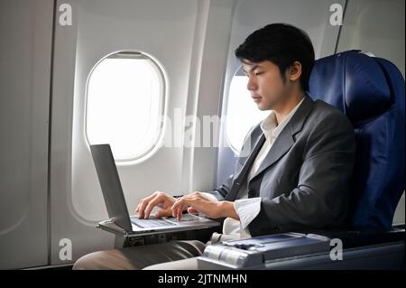 Uomo d'affari asiatico professionista che lavora, gestendo le sue attività aziendali su un computer portatile durante il volo per il suo viaggio d'affari. Business e. Foto Stock