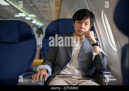 Bell'uomo d'affari asiatico e giovane astuto o capo d'affari in abito formale siede al posto della finestra in una classe business. Trasporto aereo concep Foto Stock