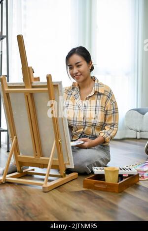 Ritratto, ispirato e talentuoso giovane artista asiatica pittura con un colore acrilico sul suo cavalletto vuoto tela nel suo salotto di casa. Foto Stock