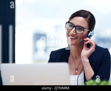 E come posso aiutarla oggi. Una donna d'affari felice che parla con la sua cuffia mentre lavora sul suo notebook in ufficio. Foto Stock