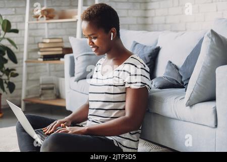 Giovane donna afro americana felice, seduta sul pavimento godendo di lavorare online sul laptop a casa. Navigare in internet, trascorrere del tempo a casa in salotto Foto Stock