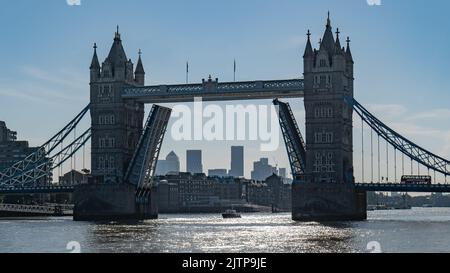 Un iconico Tower Bridge rialzato al sole del mattino con un autobus londinese in attesa di attraversare e lo skyline di Canary Wharf che si staglia tra i basculi. Foto Stock