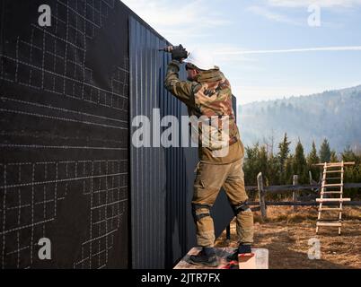 Costruttore maschio che installa nero ondulato foglio di ferro usato come facciata del cottage futuro. Uomo lavoratore edificio legno telaio casa. Carpenteria e concetto di costruzione. Foto Stock