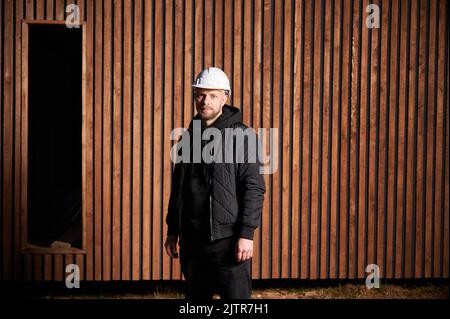 Ritratto di costruttore maschile costruzione casa con struttura in legno. Uomo in piedi sul cantiere con casco di sicurezza, su parete di fondo in legno. Foto Stock
