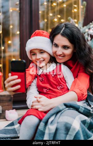Madre sorridente con figlia in cappello Santa facendo selfie via telefono mentre si siede sul portico della casa con albero di Natale decorato. ev parenting singolo Foto Stock