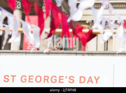 Uno spettatore è visto dietro le decorazioni mentre le persone si riuniscono per le celebrazioni del giorno di San Giorgio a Trafalgar Square, nel centro di Londra. Foto Stock