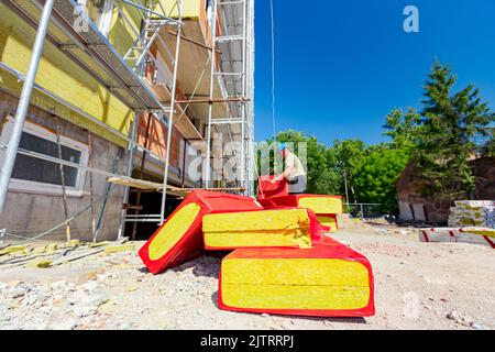 La pila di materiale termoisolante è avvolta con un foglio di cellofan. Lavoratore appende un pacchetto con lana di roccia sul gancio della gru, che lo solleva Foto Stock