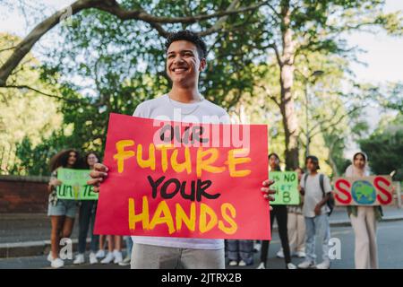 Ragazzo giovane allegro che guarda lontano mentre tiene un cartello di bandiera durante un rally cambiamento climatico. Felice giovane attivista che protesta contro il riscaldamento globale wi Foto Stock