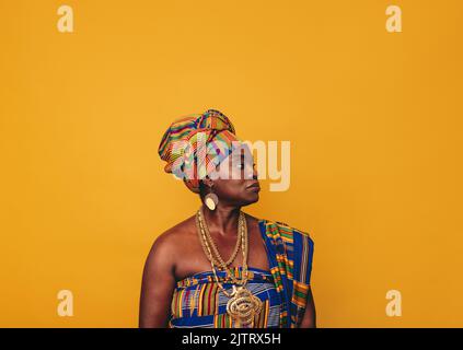Donna africana che indossa abiti tradizionali eleganti su sfondo giallo. Donna matura nera vestita di tessuto colorato Kente e gioielli d'oro Foto Stock