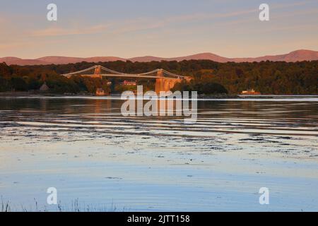 Il ponte sospeso Menai è immerso in una calda luce serale con le montagne Snowdonia sullo sfondo. Anglesey, Galles del Nord, Regno Unito. Foto Stock