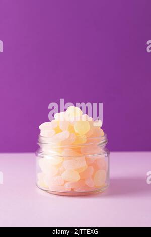 Primo piano di caramelle di gelatina in vaso di vetro su sfondo viola. Vista frontale, messa a fuoco selettiva, spazio di copia. Foto Stock