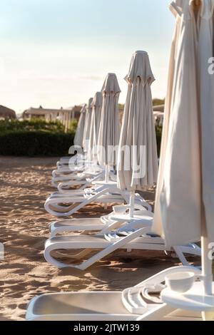 File di ombrelloni ripiegati e lettini vuoti sullo sfondo di sabbia. Resort hotel. Concetto di fine stagione turistica. Foto Stock