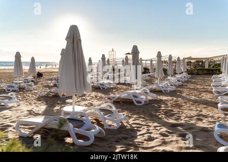 File di ombrelloni ripiegati e lettini vuoti sullo sfondo di sabbia e mare. Resort hotel. Concetto di fine stagione turistica. Foto Stock