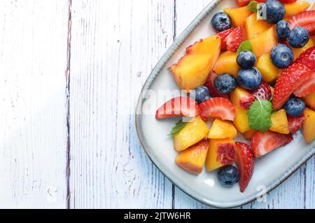 Macedonia di frutta in un piatto rettangolare su fondo di legno bianco. Fragole, pesche e mirtilli a fette con foglie di menta. Foto Stock