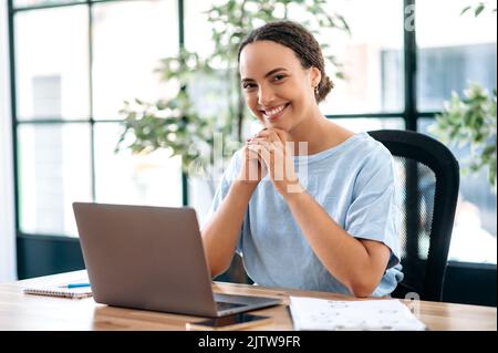 Ritratto di successo sicuro bella donna brasiliana o ispanica business, impiegato in ufficio, indossando elegante abbigliamento formale, seduto sul posto di lavoro in ufficio moderno, guardando la fotocamera, sorridendo Foto Stock