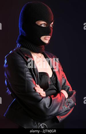 Giovane donna in balaclava e giacca in pelle in piedi con mani incrociate su sfondo nero Foto Stock