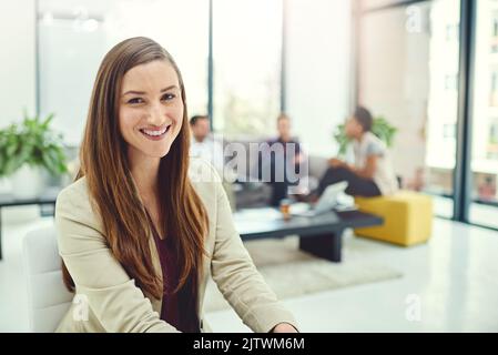 Mantenere una visione chiara dei suoi obiettivi. Ritratto di una giovane donna d'affari fiduciosa seduta in ufficio con colleghi sullo sfondo. Foto Stock