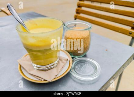 Caffè giallo dolce, servito in un bicchiere, con un cucchiaio dopo essere stato mescolato con zucchero demerara, posto all'aperto su un tavolo da ristorante. Foto Stock