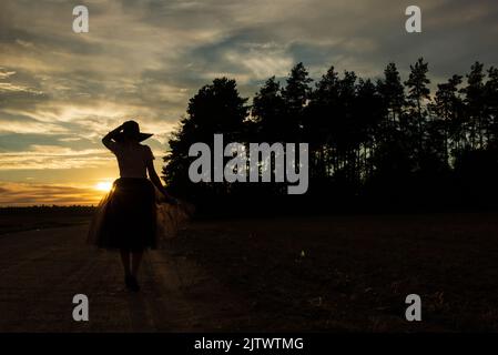 Silhouette di donna che tiene il cappello e bordo di gonna tulle camminando lungo la strada di campagna dopo il tramonto del sole contro lo skyline della foresta e cielo nuvoloso Foto Stock