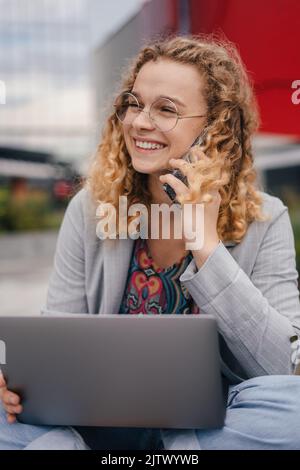 Giovane donna entusiasta ricci-haired studente in giacca grigia seduto di fronte agli edifici, resto utilizzando il portatile parlare dal telefono cellulare. Estate, internet Foto Stock
