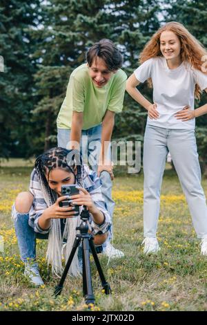 Due amici che guardano una ragazza che sta configurando lo smartphone su un terreno di gioco all'aperto con cavalletto per creare contenuti per i social media nel parco estivo. Internet Foto Stock