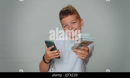 Rich toddler ragazzo guardare smartphone visualizzare sinceramente gioire vittoria, ricevendo denaro in dollari banconote in contanti, successo lotteria fortuna. Bambini ragazzi ragazzi bambini bambini piccoli isolato su sfondo grigio studio Foto Stock