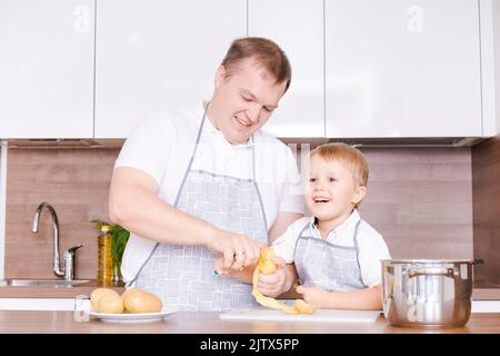 Concetto di cibo e nutrizione. Foto di due allegri papà e figlio che si posano in cucina peeling patate andando a cucinare cibo vegetariano per cena insieme, indossando grembiuli identici Foto Stock