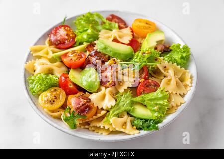 Insalata di farfalle di pasta con pomodoro, pancetta, avocado e senape condimento in un piatto su tavolo bianco. Primo piano. Cibo italiano sano Foto Stock