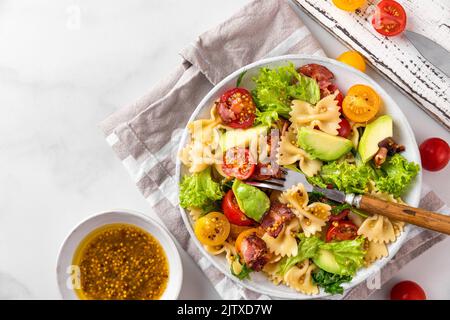 Deliziosa insalata italiana a base di farfalle di pasta, pomodori, avocado, pancetta e senape in un piatto con forchetta su tavola bianca. Vista dall'alto. Cibo sano Foto Stock