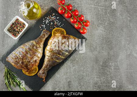 Pesce grigliato con limone, rosmarino, pomodori, olio d'oliva e spezie su piatto nero d'ardesia, su fondo grigio. Vista dall'alto, disposizione piatta con copia Foto Stock