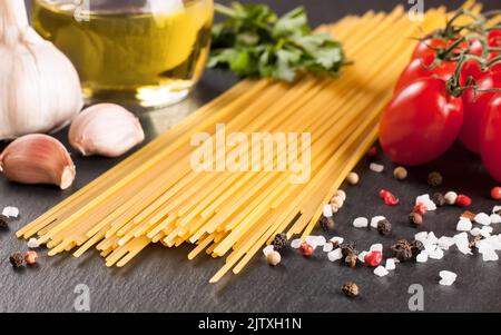 Pasta e ingridienti di cottura su superficie di ardesia nera. Spaghetti, pomodoro, olio d'oliva, aglio, prezzemolo e spezie. Cucina italiana Foto Stock