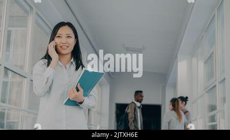 Ritratto di giovane bella studentessa di etnia asiatica che parla al telefono sorridendo positivamente e ridendo tenendo grande copybook in piedi nel corridoio dell'università con i suoi amici dietro. Foto Stock