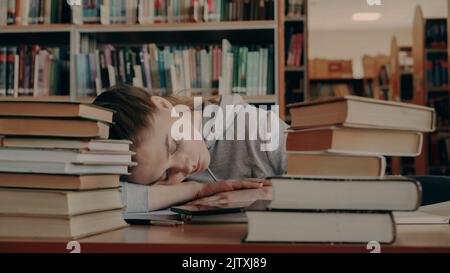 Giovane bella studentessa caucasica sta dormendo con la testa sul tavolo circondata da mucchi di libri e carte, il telefono sta mentendo davanti a lei, è in grande biblioteca universitaria Foto Stock