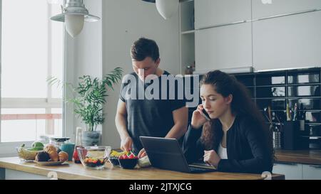 grave caucasica bella coppia in cucina, l'uomo sta tagliando l'insalata, è in piedi in silenzio, donna è seduta a tavolo di lavoro sul computer portatile e parlare al telefono Foto Stock