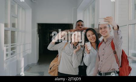 Gruppo di quattro studenti multietnici che prendono selfie su smartphone fotocamera mentre si trovano in corridoio dell'università . Hippster tipo che tiene il telefono e gli amici stanno posando positivamente Foto Stock