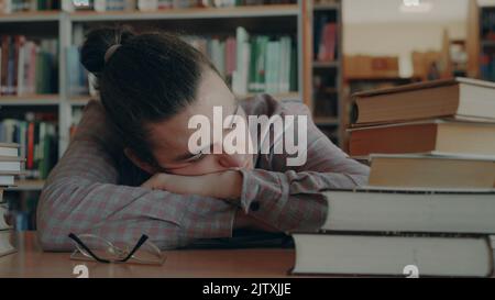 Bel giovane uomo caucasico seduto con la testa sul tavolo dorme in una grande biblioteca scolastica circondata da enormi mucchi di libri. I suoi occhiali stanno mentendo davanti a lui Foto Stock