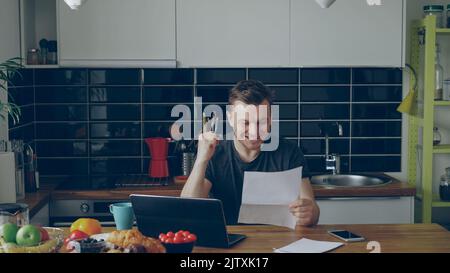 Bel giovane uomo riceve buone notizie leggendo la lettera di carta in cucina, mentre fare colazione a casa la mattina presto Foto Stock