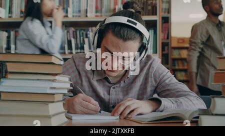 Giovane bel ragazzo caucasico seduto a tavola in grande biblioteca del college sta scrivendo qualcosa nel copybook. Sorride e sembra positivo e felice, libri sdraiati sul tavolo Foto Stock