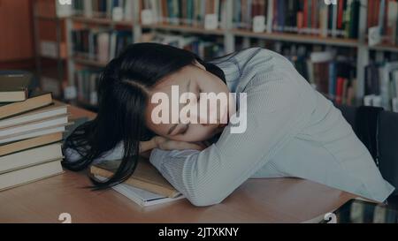 Giovane bella studentessa asiatica stanca è seduta alla scrivania con la testa su di essa, sta dormendo su libro di testo circondato da mucchi in grande biblioteca spaziosa Foto Stock