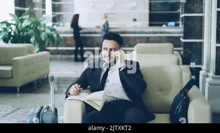 Pan shot di allegro uomo d'affari fiducioso che parla telefono cellulare sorridendo e scrivendo note in blocco note mentre si siede sulla poltrona nella reception dell'hotel. Concetto di viaggi, affari e persone Foto Stock