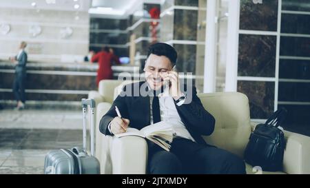 Pan shot di allegro uomo d'affari fiducioso che parla telefono cellulare sorridendo e scrivendo note in blocco note mentre si siede sulla poltrona nella reception dell'hotel. Concetto di viaggi, affari e persone Foto Stock