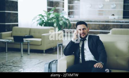 Pan shot di sicuro uomo d'affari parlando telefono cellulare mentre si siede sulla poltrona in hotel di lusso con bagagli vicino a lui. Concetto di viaggi, affari e persone Foto Stock