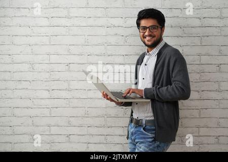 Ritratto di allegro programmatore indiano con computer portatile in piedi contro muro di mattoni bianchi Foto Stock